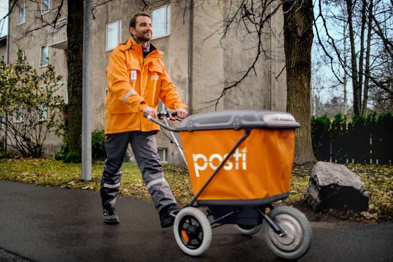 posti working in an orange rainjacket pusing an orange posti cart in the rain
